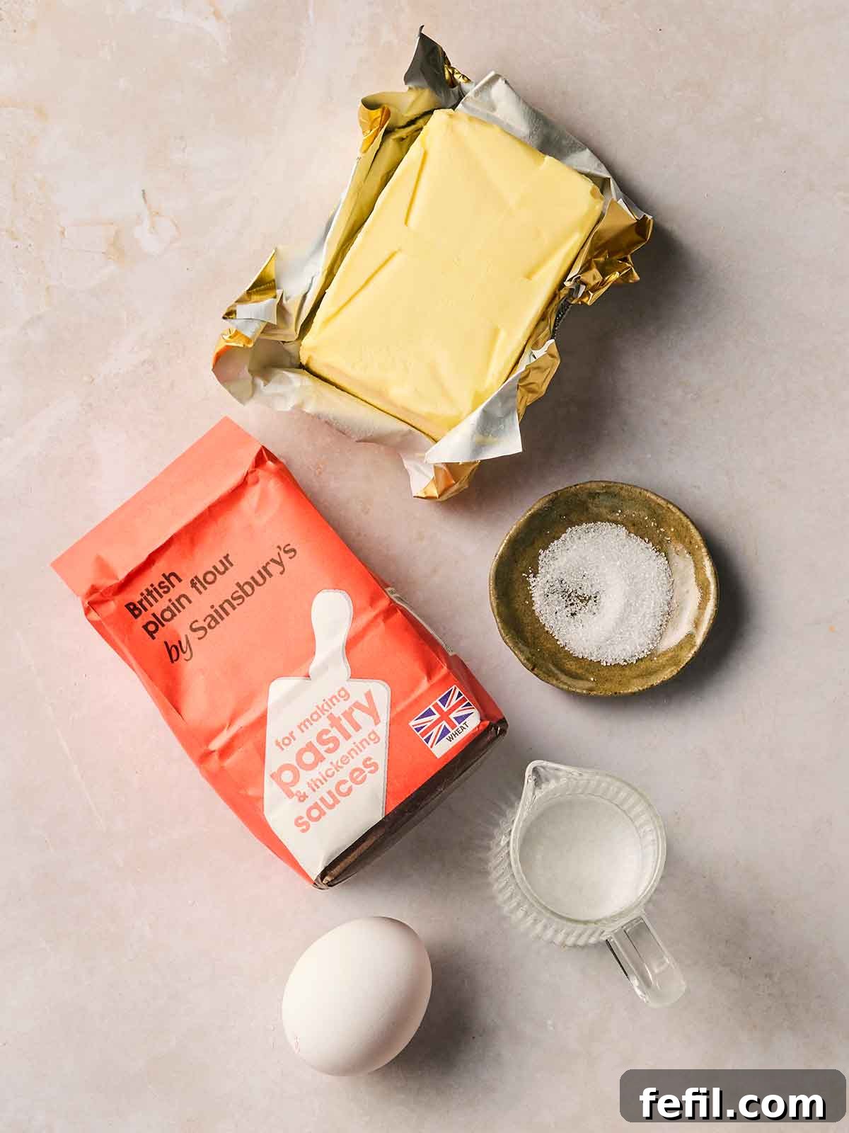 The ingredients for making shortcrust pastry on a white surface, including plain flour, butter, an egg, water and some salt.