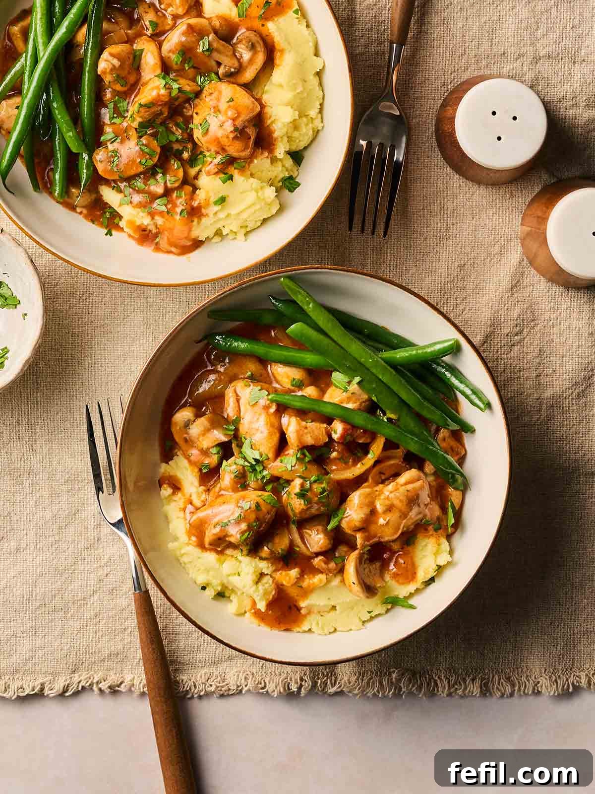 Two cozy bowls of Chicken Chasseur, garnished with fresh parsley, presented on a set table with mashed potatoes and green beans, ready for a delicious meal.