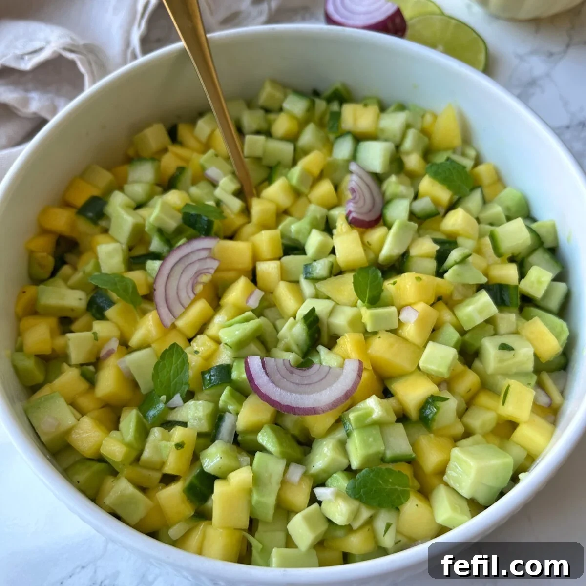 A bowl of diced mango, cucumber, and avocado salad garnished with red onion slices and mint leaves, with a gold spoon in the bowl.