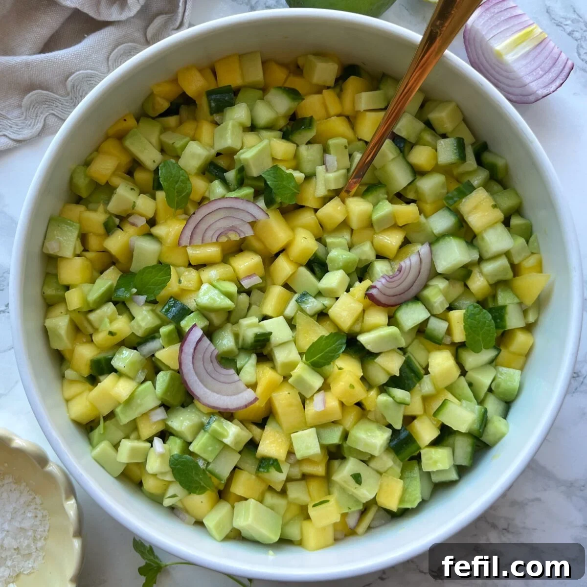 A bowl of diced mango, cucumber, and avocado salad garnished with red onion slices and mint leaves, with a serving spoon.