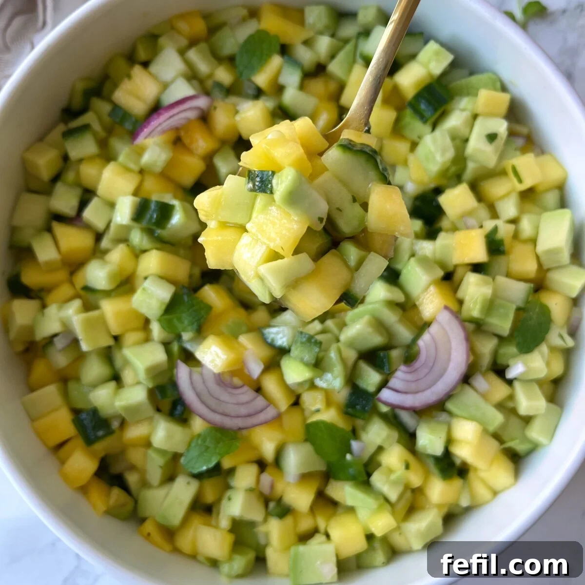 A bowl of diced mango, avocado, cucumber, red onion, and mint is displayed as a spoon lifts a portion.