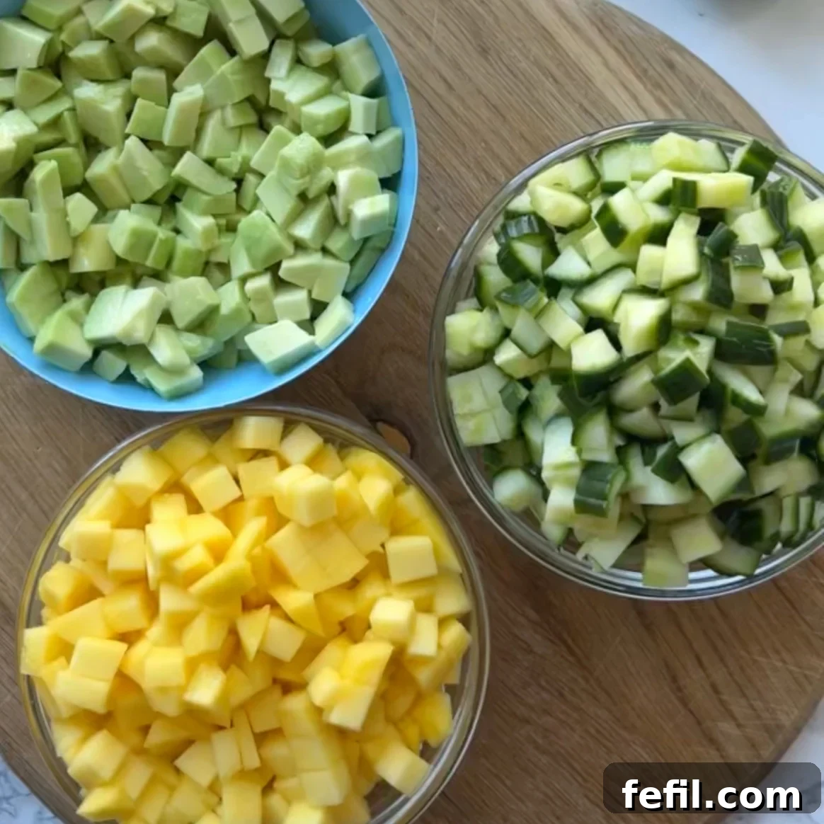Three bowls with diced mango, avocado, and cucumber are placed on a wooden board.