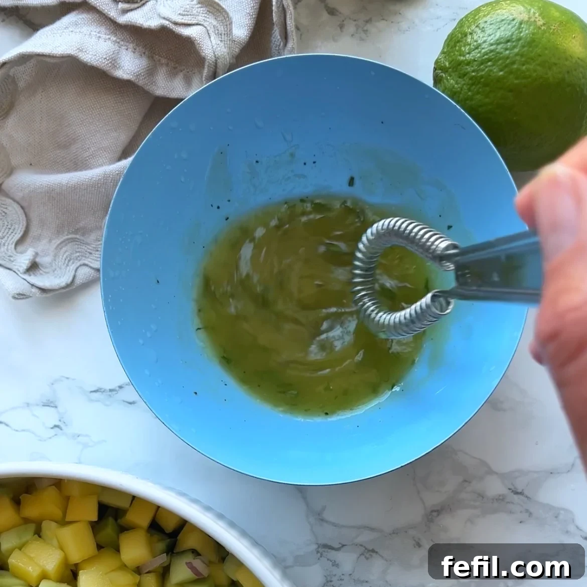 A hand whisking a green dressing in a blue bowl, with a lime, diced mango, and a cloth nearby on a marble surface.
