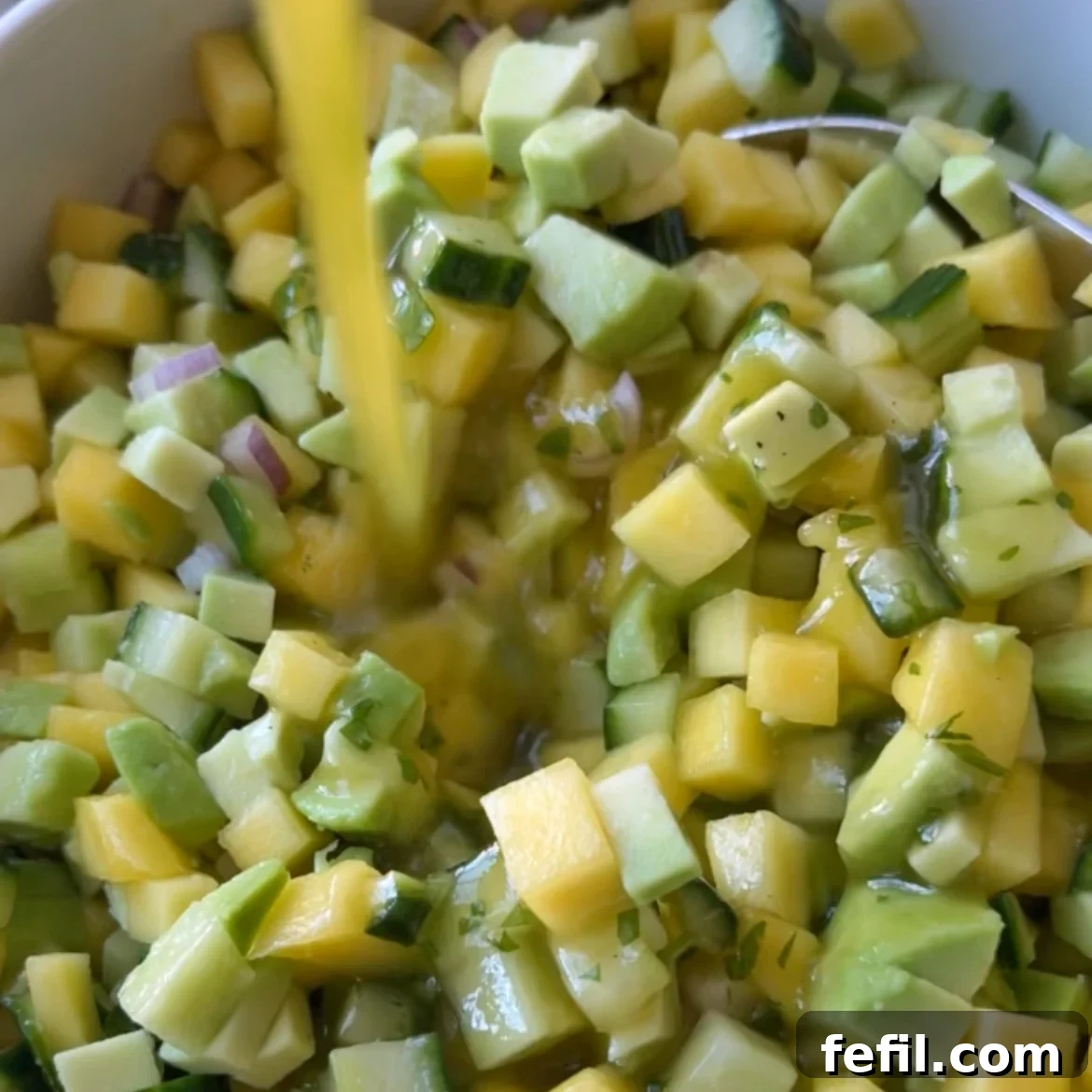 Close-up of an Avocado Mango Salad: diced avocado, mango, cucumber, and red onion in a bowl as a liquid dressing is poured over the fresh mixture.