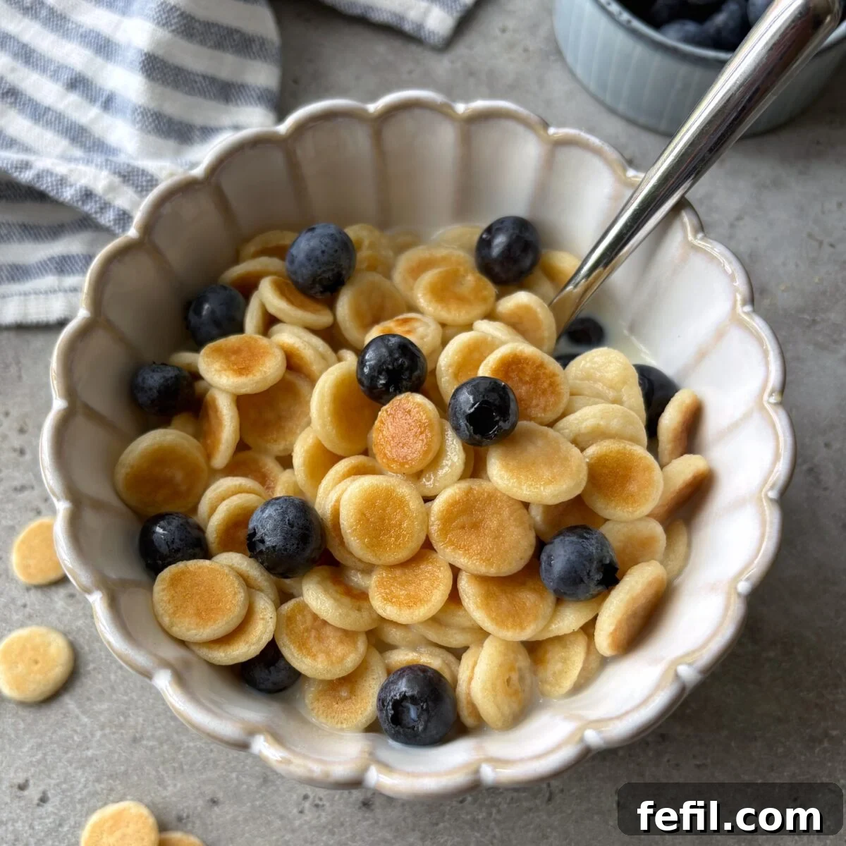 A scalloped bowl of mini pancakes topped with milk, maple syrup, and blueberries with a striped dish cloth and bowl of berries in the background.