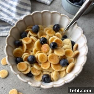 A scalloped bowl of mini pancakes topped with milk, maple syrup, and blueberries with a striped dish cloth and bowl of berries in the background.