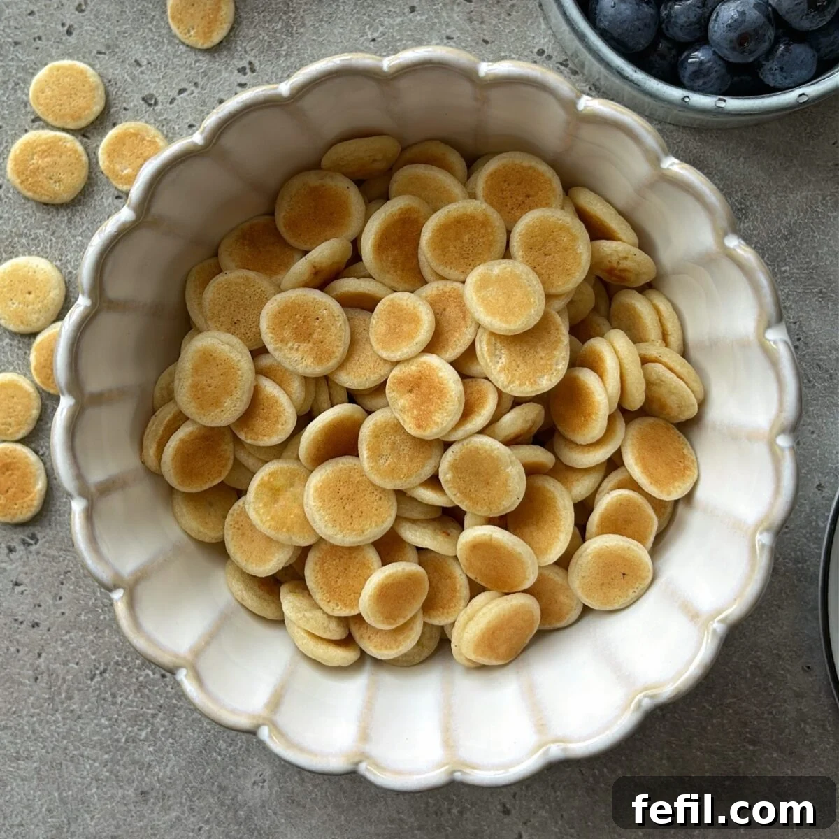 A bowl of mini pancakes ready to be topped with milk, maple syrup, and blueberries.
