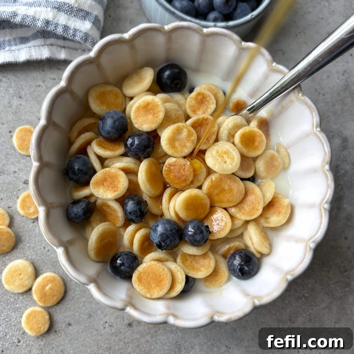 Maple syrup being poured onto a bowl of mini pancake cereal topped with blueberries and milk.