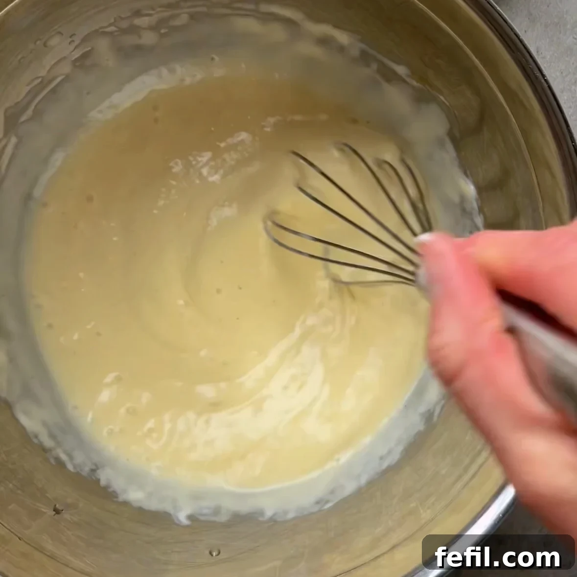 A whisk mixing pancake batter in a stainless steel bowl.