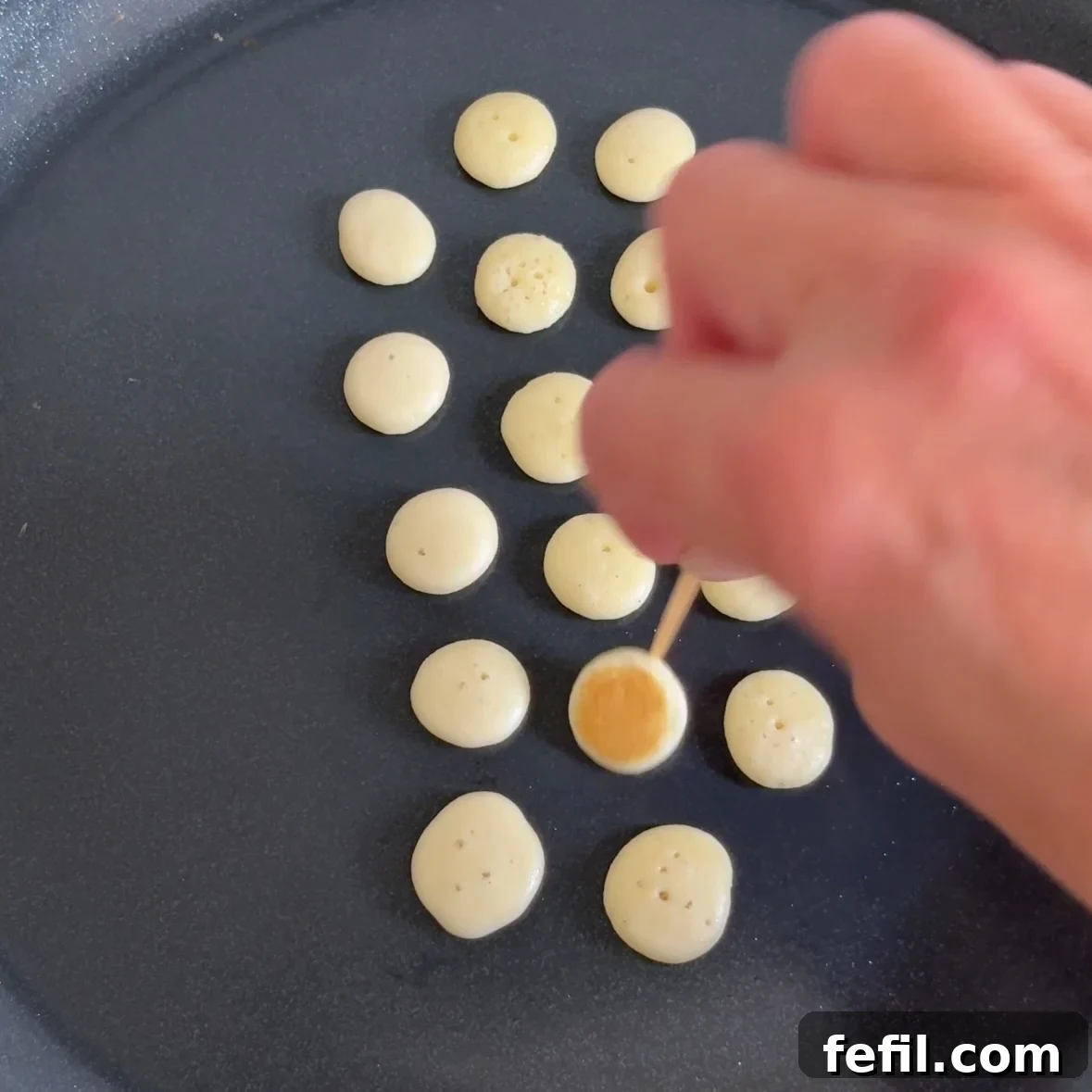 A hand uses a toothpick to turn over mini pancakes on a hot pan.