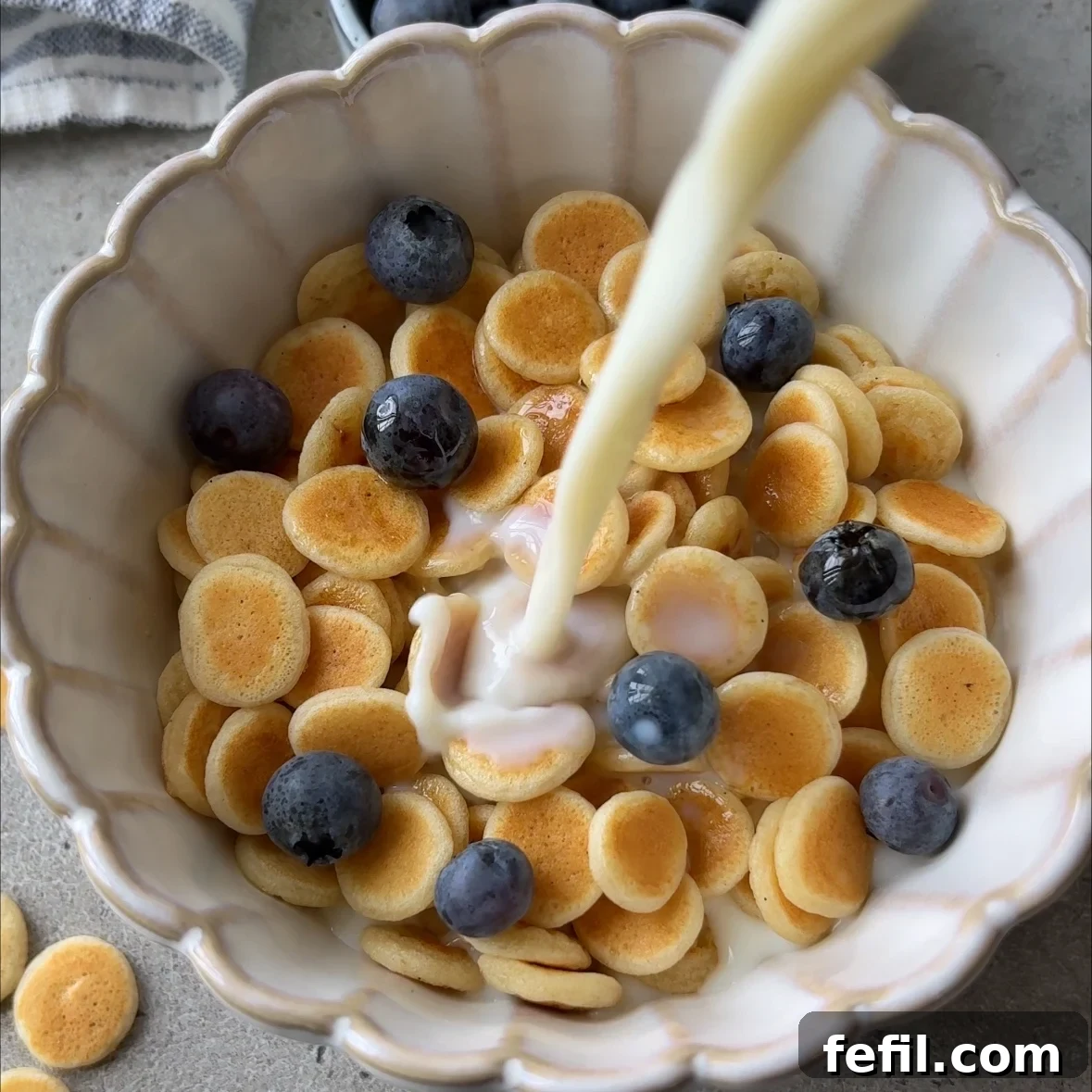 Mini pancakes in a bowl topped with maple syrup and blueberries. A hand pours milk on top.