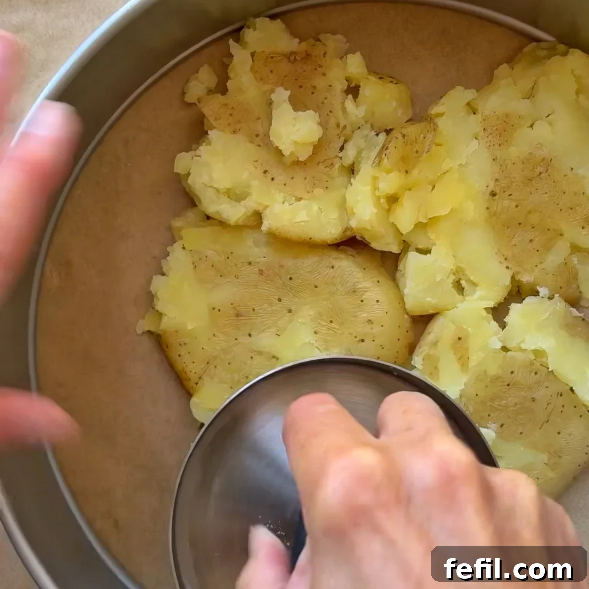 A hand smashes cooked potatoes inside a round cake pan lined with parchment.