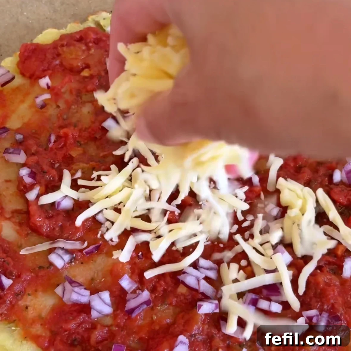 A hand sprinkles mozzarella on top of a smashed potato pizza base topped with tomato sauce and diced red onions.