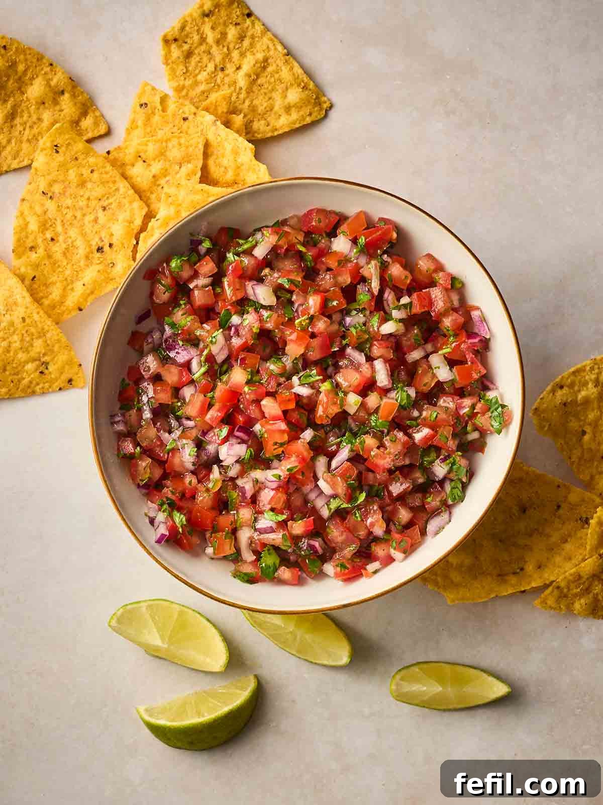 A white bowl of homemade Salsa on a white surface, with tortilla chips and lime wedges around the side.