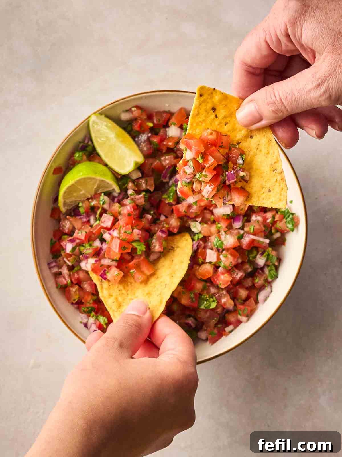 Two hands holding tortilla chips, dipping into a bowl of fresh salsa.