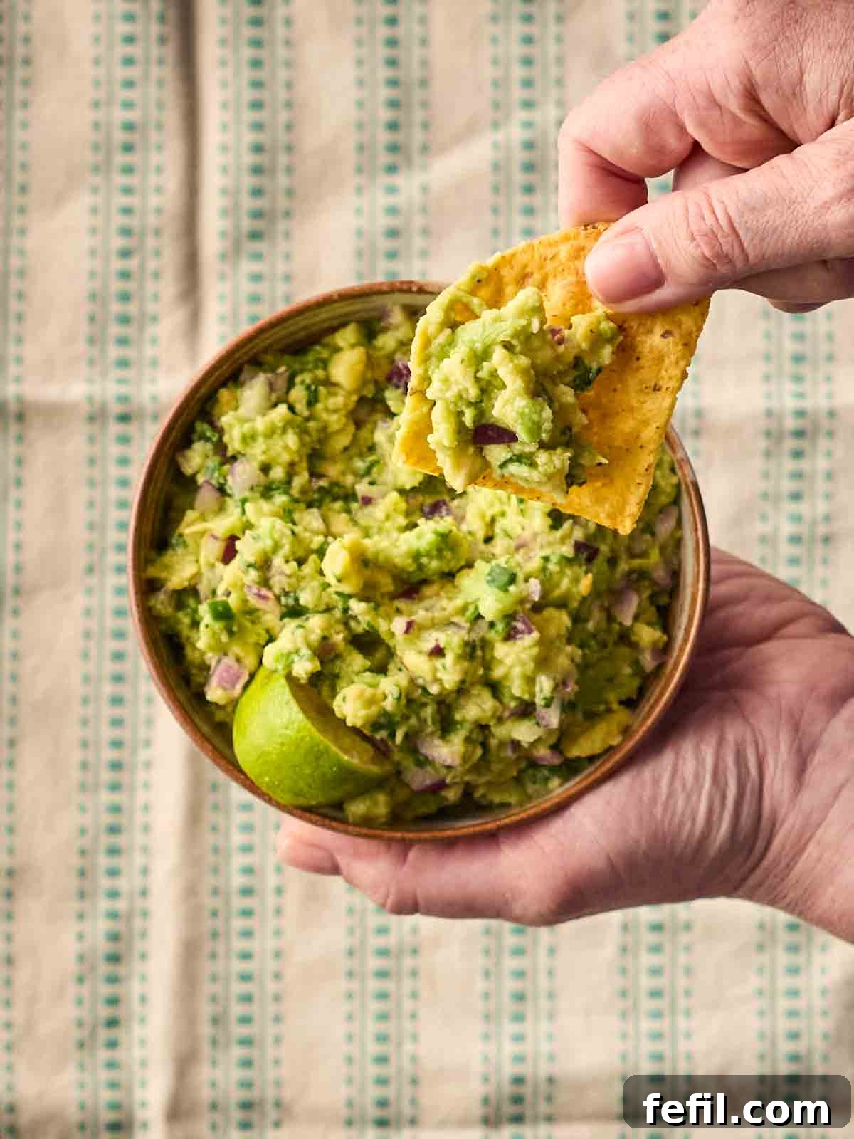 Fresh Guacamole Recipe: Zesty Homemade Mexican Dip 3 A hand holding a bowl of guacamole with another hand dipping in a tortilla chip.