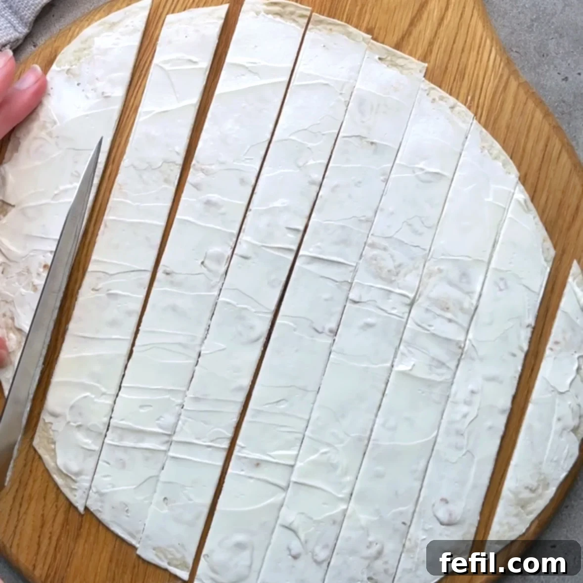 Smoked Salmon Pinwheels with Cream Cheese and Dill 4 A tortilla topped with cream cheese is being cut into strips with a knife.