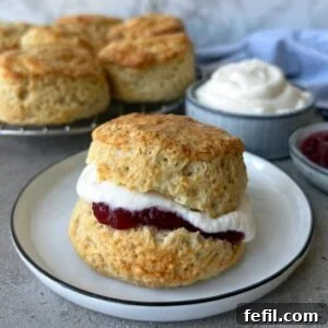 A closeup of a buttermilk scone topped with whipped cream and jam. In the background are more scones resting on a baking rack.