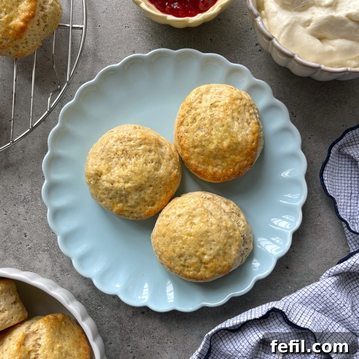 Three plain scones are arranged on a light blue scalloped plate, with cream, jam, and a cooling rack nearby.