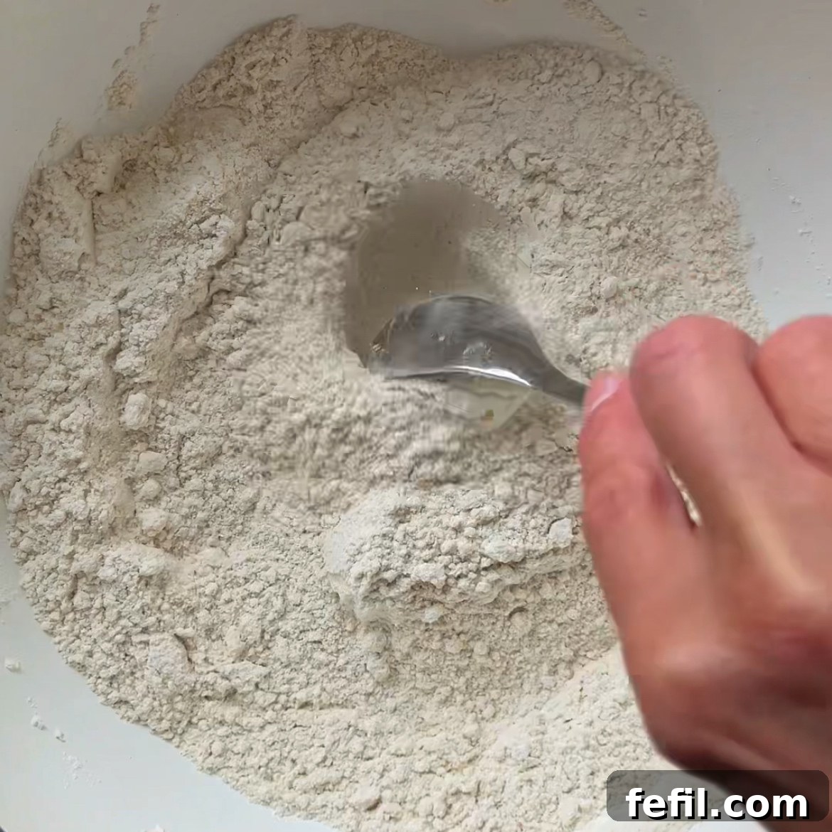 A hand mixes flour, sugar, and baking powder in a medium bowl.
