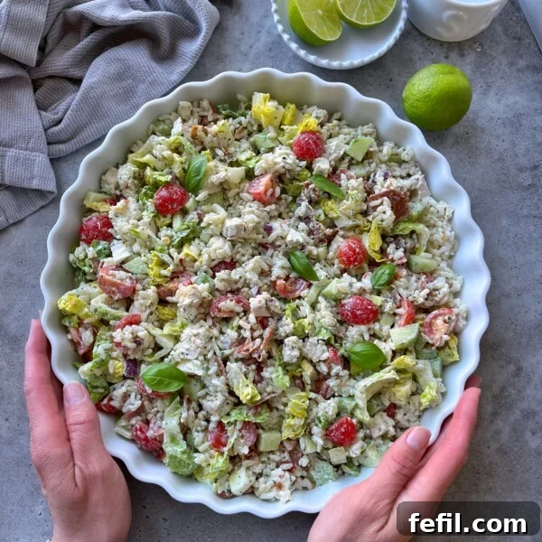 A person holds a round dish filled with a colorful rice chicken salad made of lettuce, cherry tomatoes, avocado, and fresh basil.