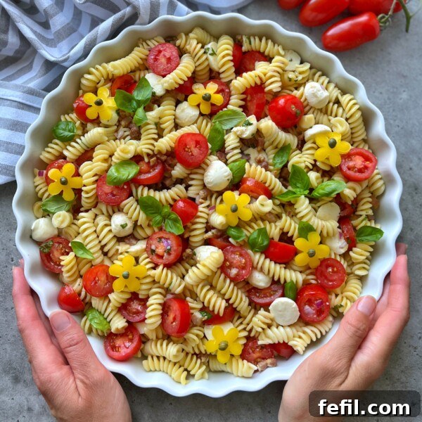 A person holds a round dish filled with healthy fusilli pasta salad, cherry tomatoes, mozzarella balls, basil leaves, and yellow flower-shaped bites.