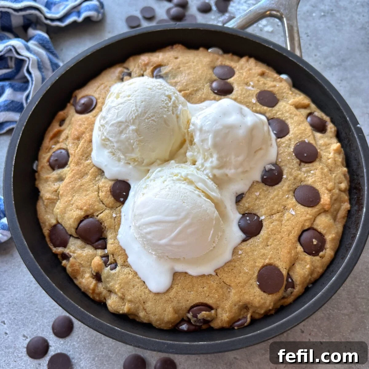 Gooey Chocolate Chip Skillet Cookie Recipe 2 Chocolate Chip skillet cookie topped with three scoops of vanilla ice cream. In the background are more chocolate chips and a blue striped tea towel.