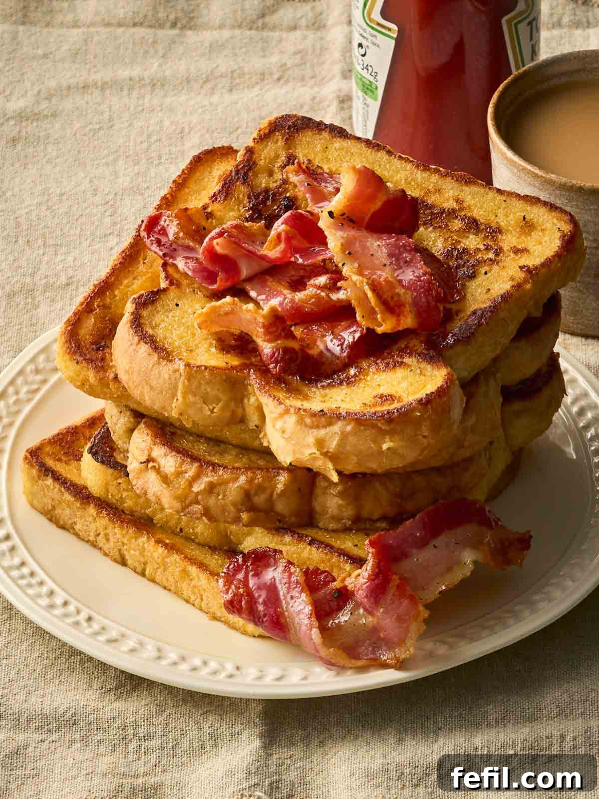 How to Make Classic Eggy Bread (British-Style French Toast) 3 A side angle of a stack of Eggy Bread with bacon on a plate, with a cup of tea in the background.