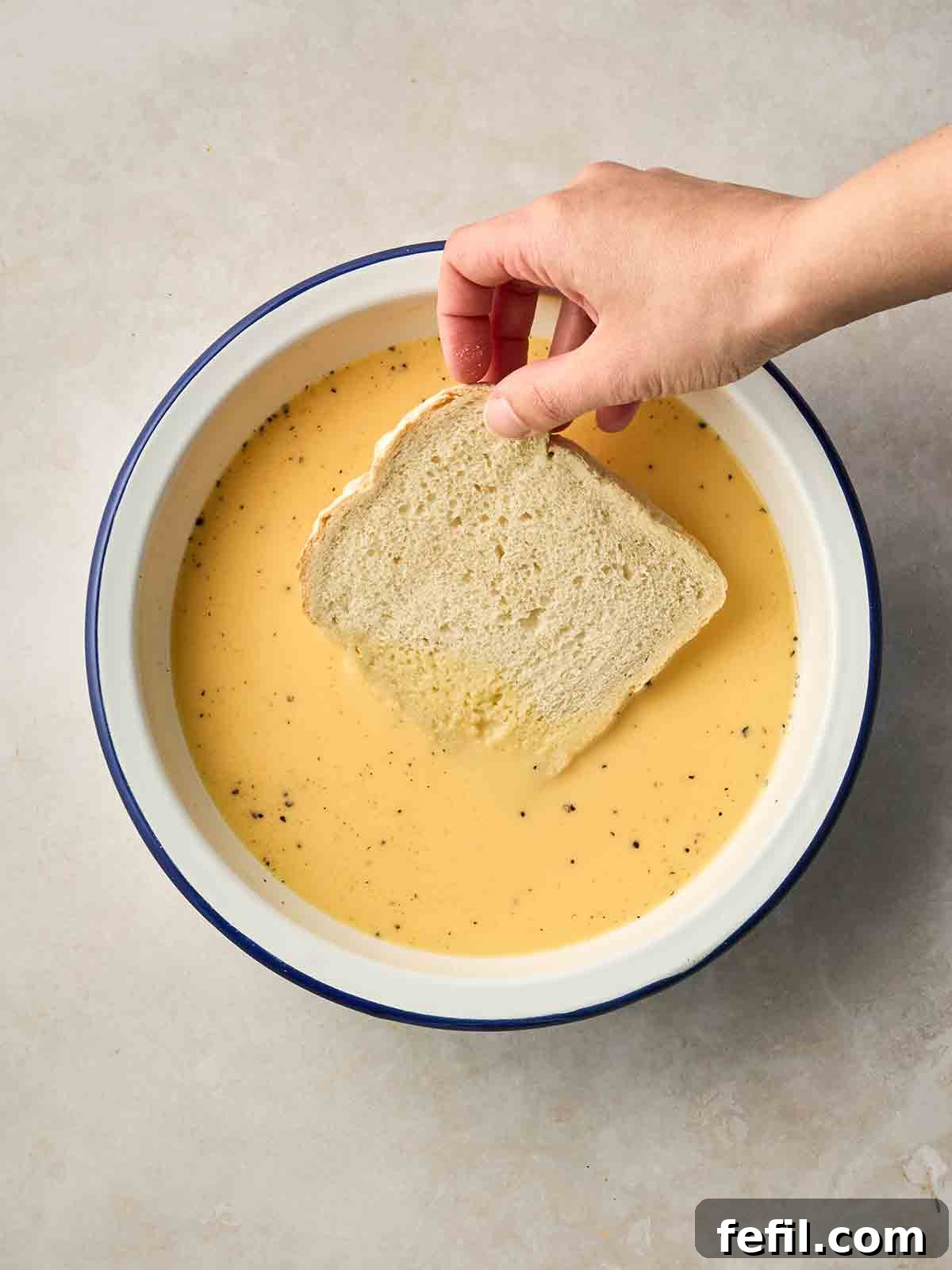 How to Make Classic Eggy Bread (British-Style French Toast) 7 A hand placing a slice of white bread into an eggy mixture.