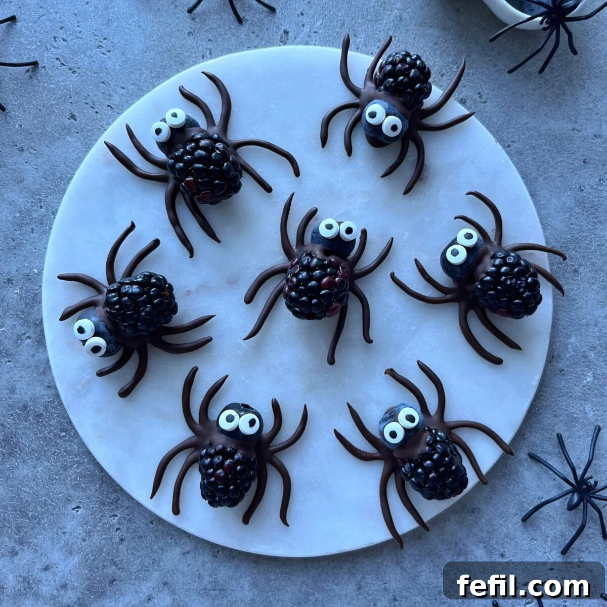 Halloween Chocolate Spider Treats for Parties 2 Seven spider-shaped treats made from blackberries, blueberries, dark chocolate, and candy eyes are arranged on a round white plate.