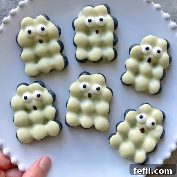 Halloween Chocolate Spider Treats for Parties 11 Six white chocolate blueberry bites shaped like bubbly ghosts, each with cute, surprised facial expressions and wide eyes, are arranged on a scalloped-edge white plate. A hand holds the edge the plate.