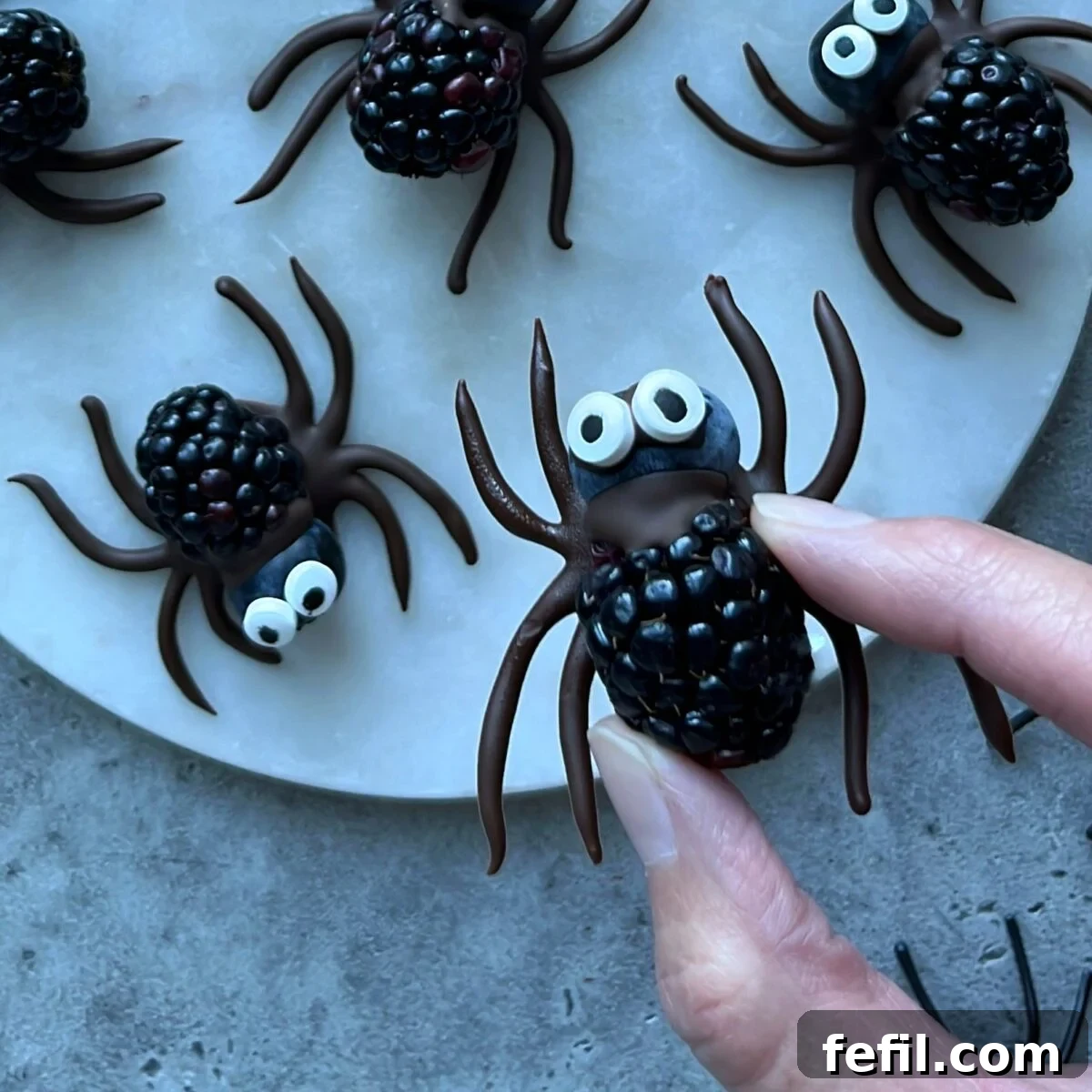 Halloween Chocolate Spider Treats for Parties 3 Hand holding a blackberry decorated as a spider with a blueberry head, chocolate legs and candy eyes, with similar blackberry spiders arranged on a white plate.