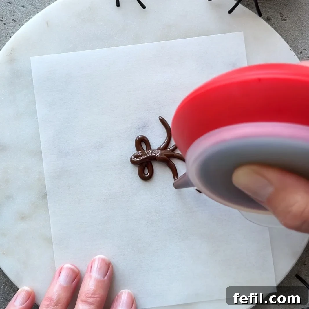 Halloween Chocolate Spider Treats for Parties 4 A hand uses a red piping tool to pipe chocolate onto parchment paper.