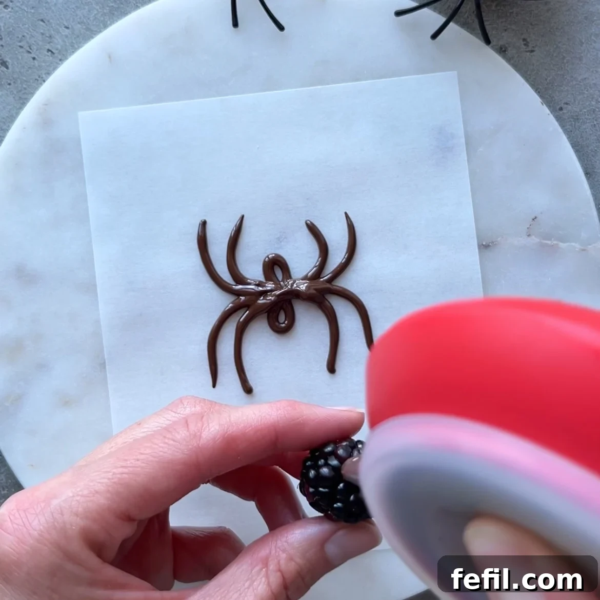 Halloween Chocolate Spider Treats for Parties 5 A hand pipes chocolate onto a blackberry above a sheet of parchment featuring delicious Chocolate Spiders.
