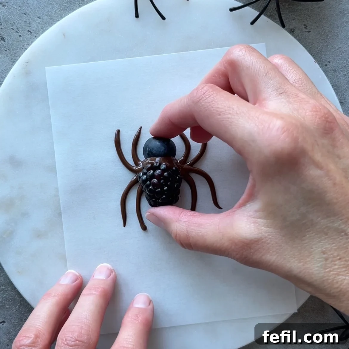 Halloween Chocolate Spider Treats for Parties 6 A hand places a blackberry and a blueberry onto a chocolate spider piped on parchment paper.