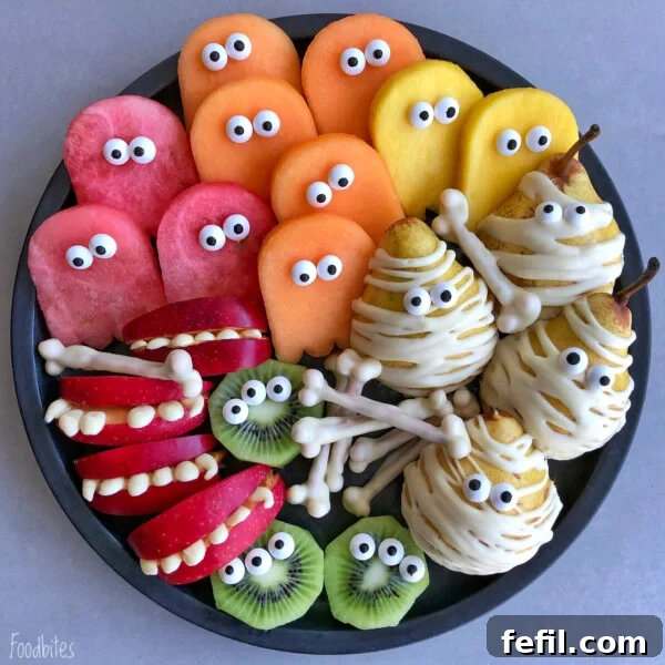 Halloween Chocolate Spider Treats for Parties 8 A plate of Halloween-themed bites features apple slices with teeth, kiwi slices, fruit cutouts with candy eyes, and pears wrapped like mummies with white chocolate and candy eyes.