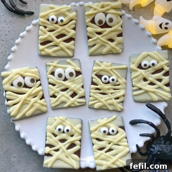 Halloween Chocolate Spider Treats for Parties 9 A plate of rectangular chocolate pieces decorated as mummies with white chocolate bandages and candy eyes. Plastic spider and ghost decorations surround the plate.