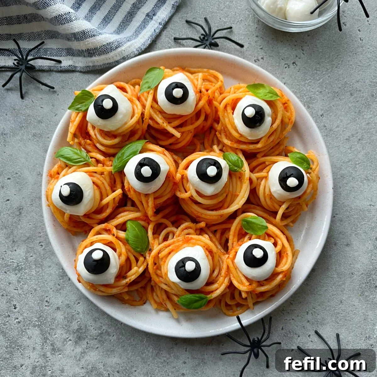 Spooky Halloween Spaghetti with Creepy Meatball Eyes 2 A plate of spaghetti nests topped with mozzarella balls, sliced olives, and basil leaves, arranged to look like eyeballs. Plastic spiders and a striped napkin are nearby.