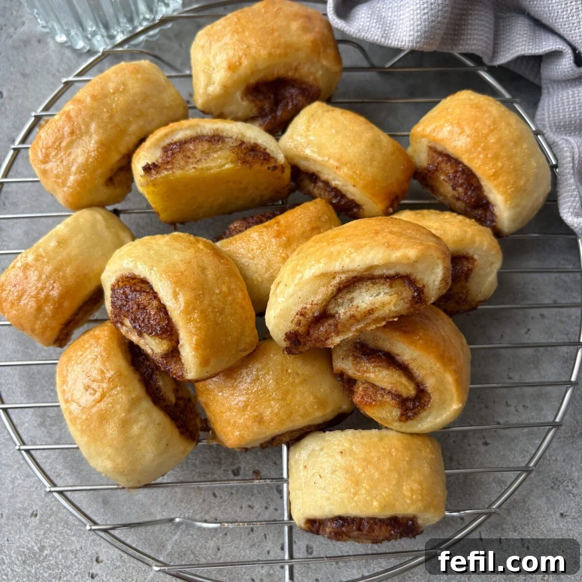 Mini Cinnamon Roll Bites with Cream Cheese Glaze 3 Golden brown mini cinnamon rolls cooling on a round metal rack, with a grey cloth in the background.