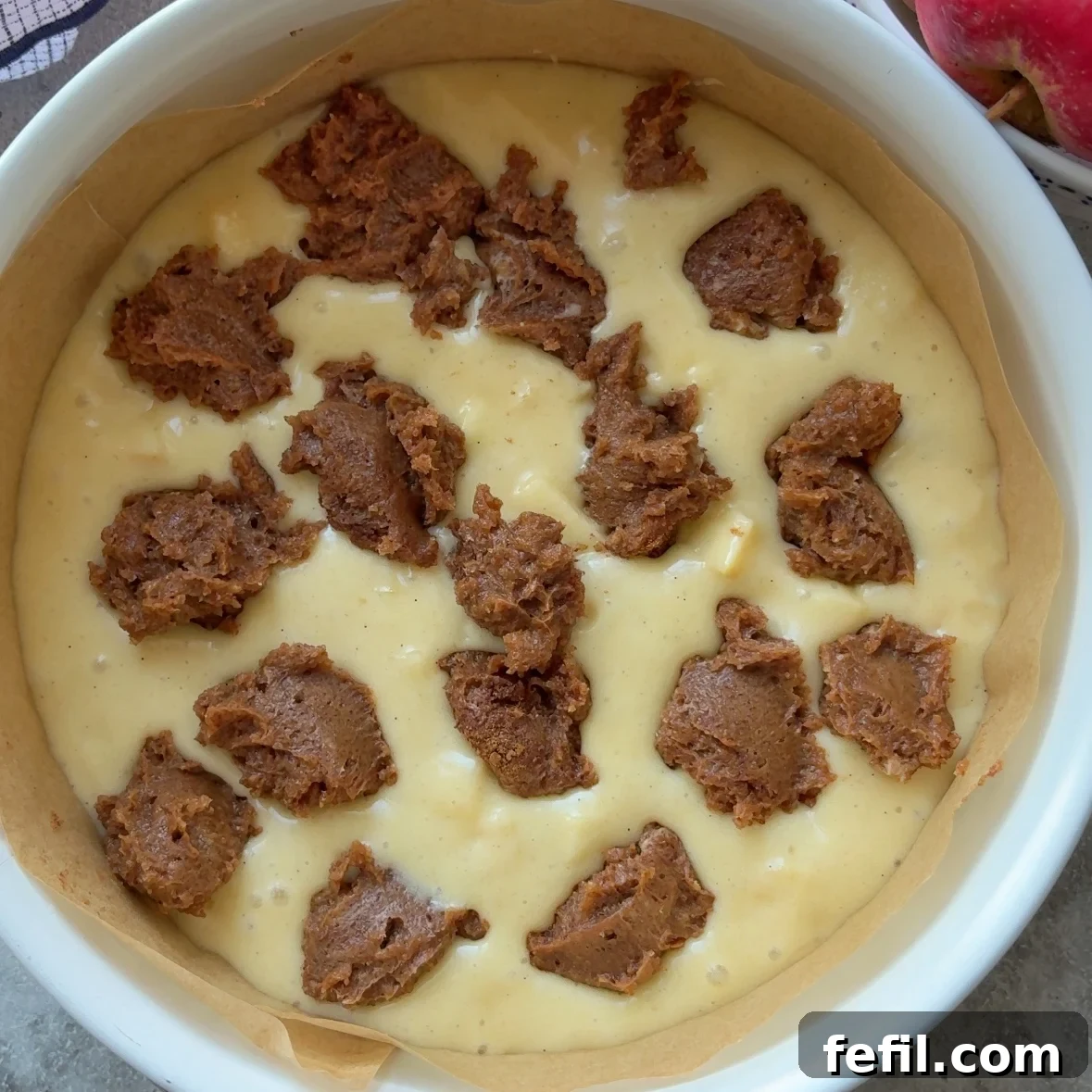 Classic Cinnamon Apple Cake with Warm Spice Glaze 11 A round baking dish lined with parchment paper contains pale yellow batter with uneven chunks of brown sugar filling scattered on top, ready to become a delicious Cinnamon Apple Cake.