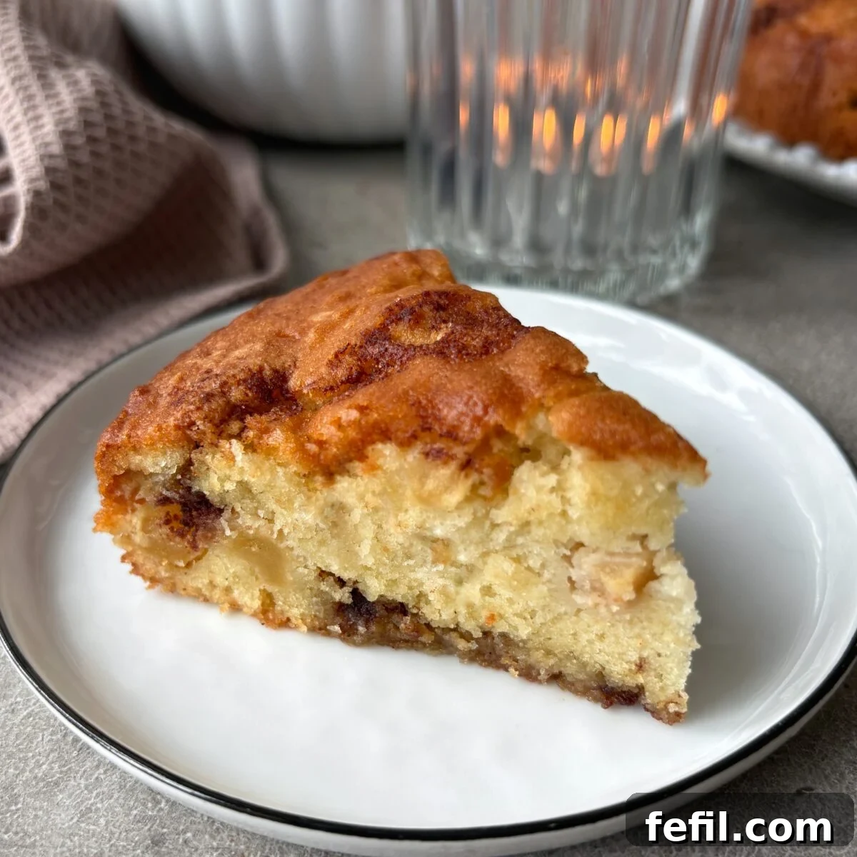 Classic Cinnamon Apple Cake with Warm Spice Glaze 3 A slice of Cinnamon Apple Cake on a small white plate with a kitchen towel and a tealight in the background.