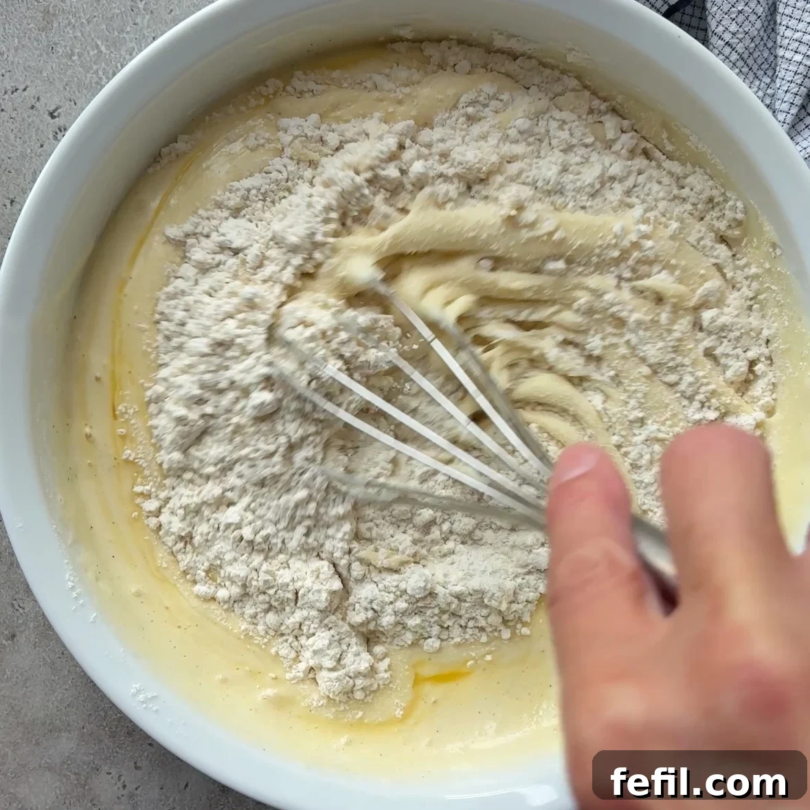 Classic Cinnamon Apple Cake with Warm Spice Glaze 9 A hand uses a whisk to mix flour into a yellow batter for Cinnamon Apple Cake in a white bowl, with a kitchen towel partially visible in the corner.