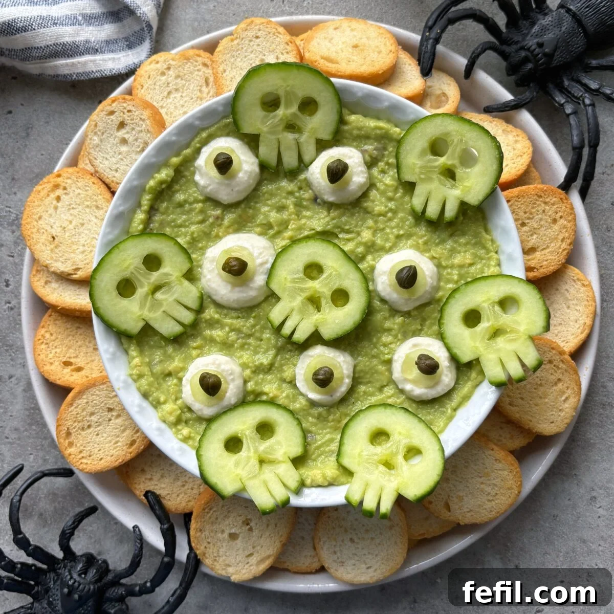 Spooky Halloween Guacamole Recipe for Party Appetizers 2 A bowl of green dip topped with cucumber skulls and surrounded by toasted bread slices, with plastic spider decorations nearby.