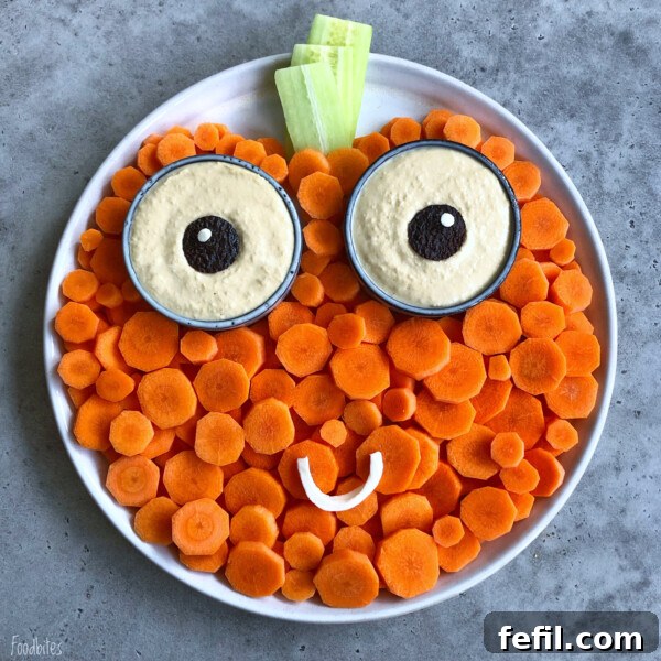 A round plate arranged with carrot slices, two cups of hummus topped with olives for eyes, cucumber pieces for a stem, and a curved vegetable smile creates a pumpkin face.