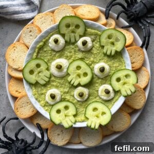 A bowl of green dip topped with cucumber skulls and surrounded by toasted bread slices, with plastic spider decorations nearby.