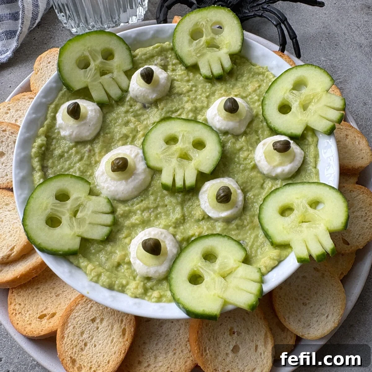 Spooky Halloween Guacamole Recipe for Party Appetizers 3 A sideview of a bowl of guacamole topped with cucumber slices cut into skull shapes, dollops of yogurt dip and capers, surrounded by round toast pieces.
