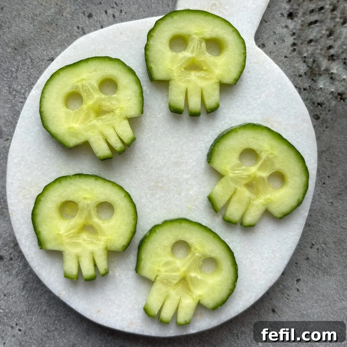 Spooky Halloween Guacamole Recipe for Party Appetizers 4 Five cucumber slices cut to resemble skull shapes are arranged on a round white cutting board placed on a gray surface.