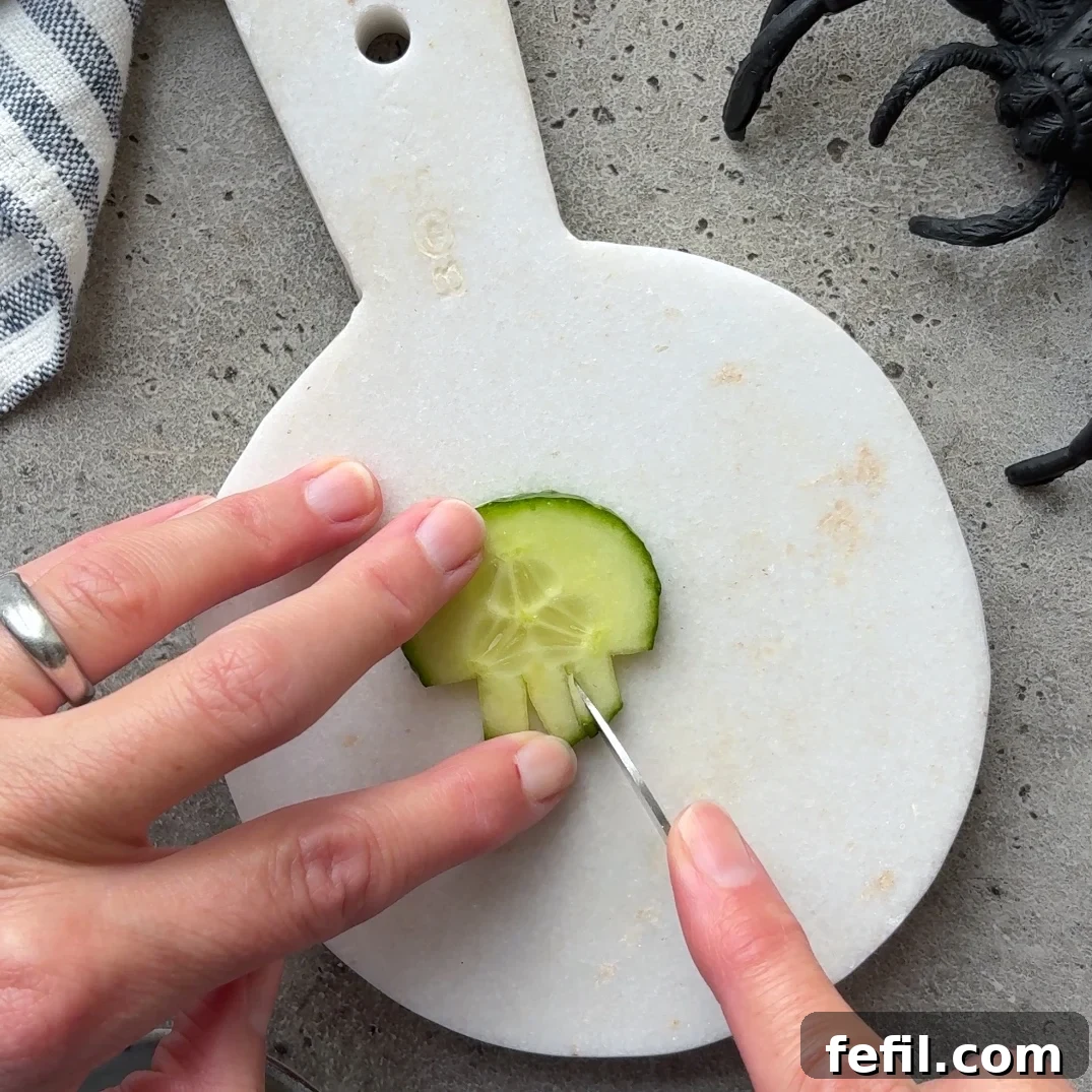 Spooky Halloween Guacamole Recipe for Party Appetizers 7 A hand carves a cucumber slice into a skull shape on a white cutting board.