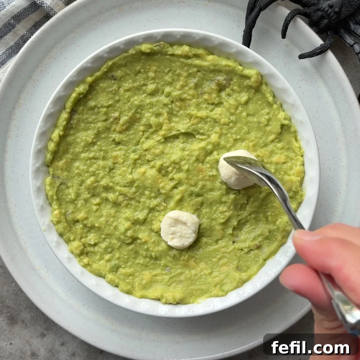 Spooky Halloween Guacamole Recipe for Party Appetizers 8 A hand uses a spoon to shape two round dollops of garlic dip into eyeballs on top of a bowl of Halloween Guacamole, with a toy spider partially visible in the background.