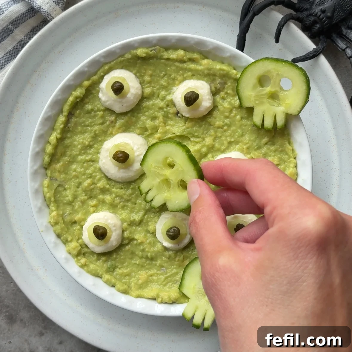 Spooky Halloween Guacamole Recipe for Party Appetizers 9 A hand places a cucumber skull on Halloween Guacamole topped with garlic dip eyeballs on a white plate with a plastic spider nearby.